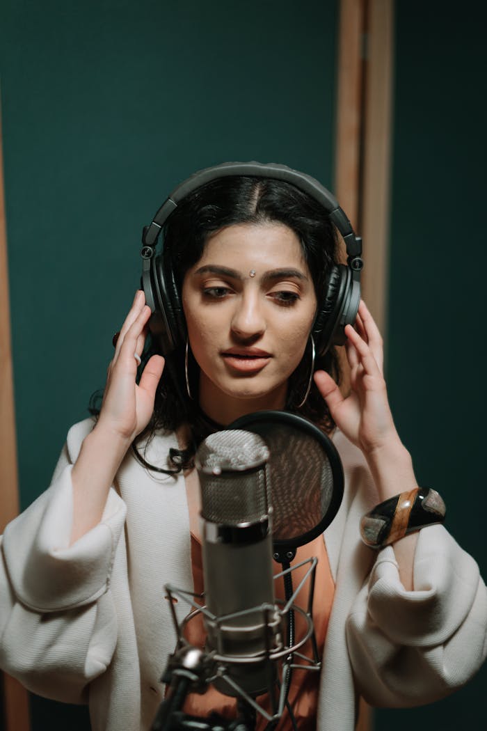 A young woman sings into a microphone in a professional music studio setting, wearing headphones.